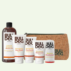 The Energising Collection Bundle includes shower gel, face wash, face scrub, moisturizer, and natural deodorant, displayed in front of a brown zippered toiletry bag with white and brown packaging featuring orange accents.