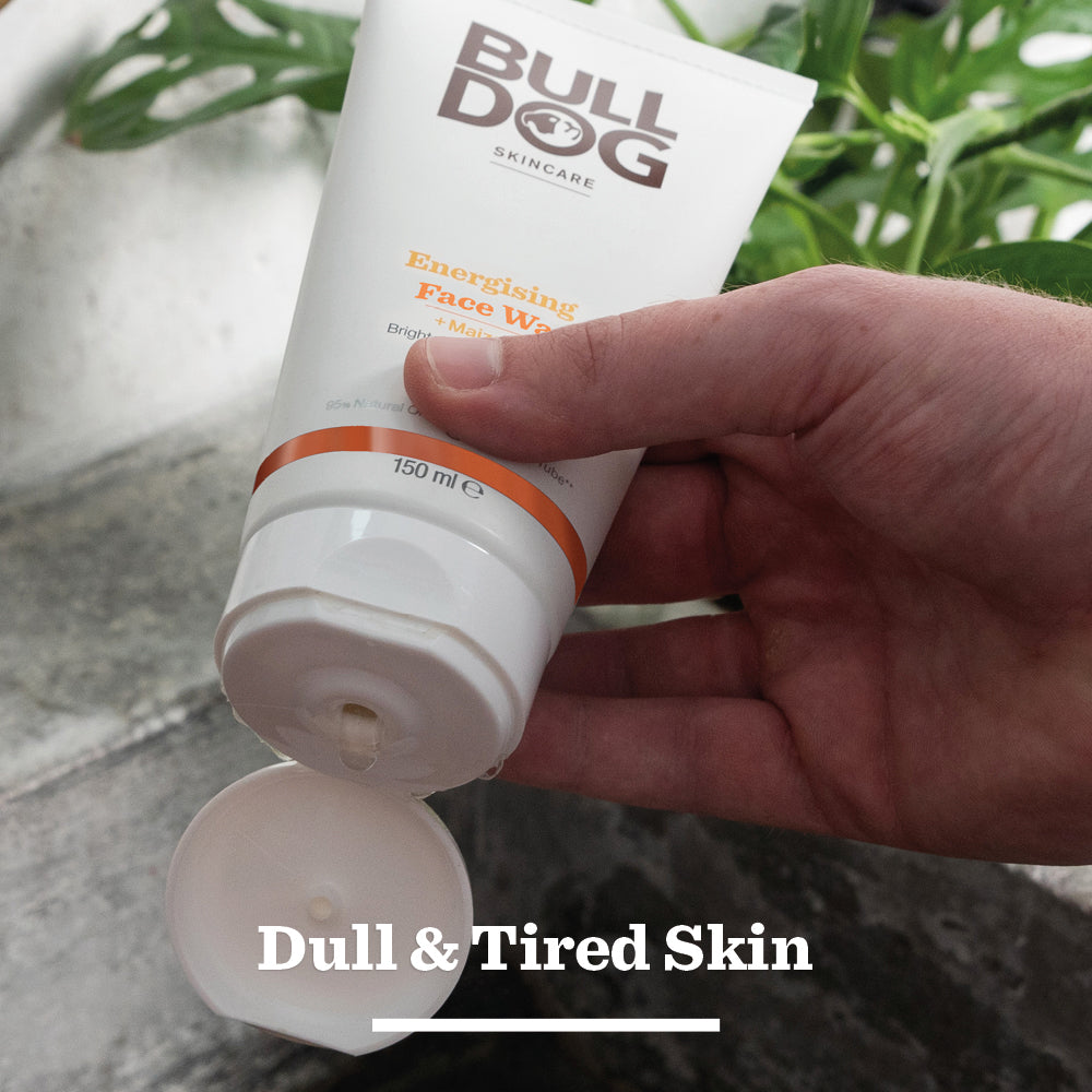 A hand squeezes a tube of Bulldog Skincare Face Wash over a sink, with “Dull & Tired Skin” text below. Green plants and the Energising Spray Deodorant Bundle appear in the background.