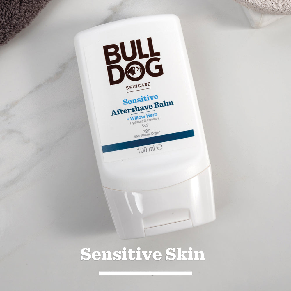 A white bottle of Sensitive Aftershave Balm rests on a white marble surface. The label highlights care for sensitive skin and natural origin ingredients like willow herb. “Sensitive Skin” is written below the bottle.