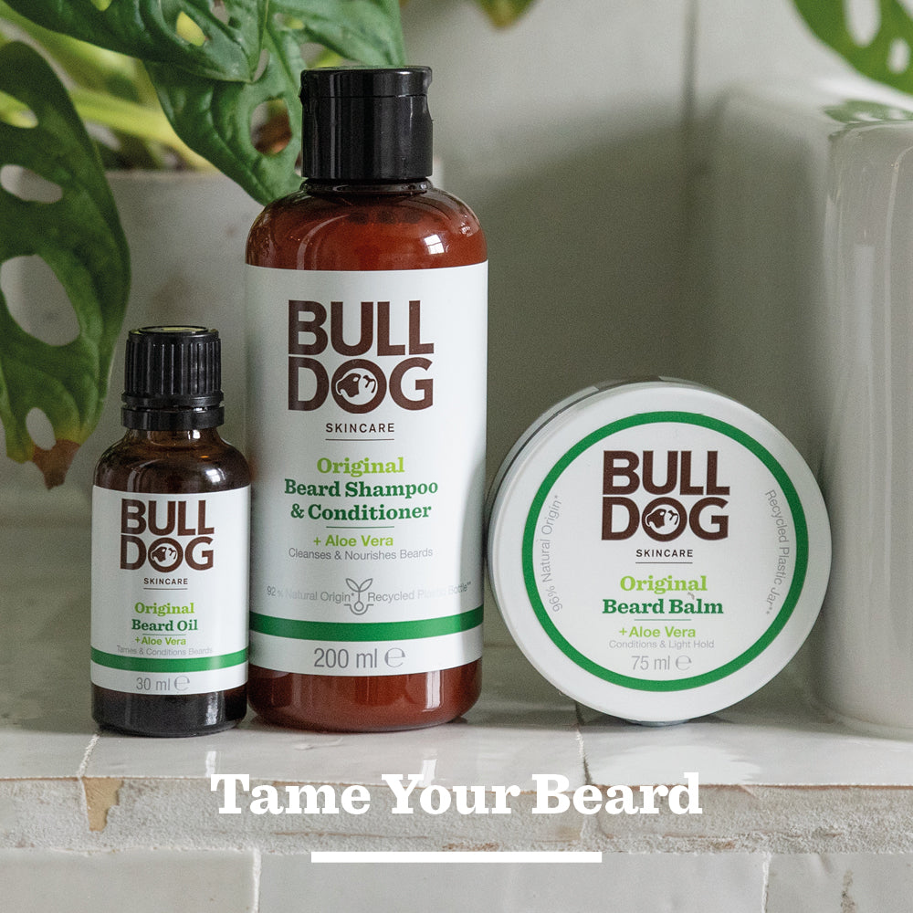 Bulldog Skincare’s Original Beard Shampoo and Conditioner is shown on a countertop with green leaves in the background. Made with natural origin ingredients. Text at the bottom reads, Tame Your Beard.