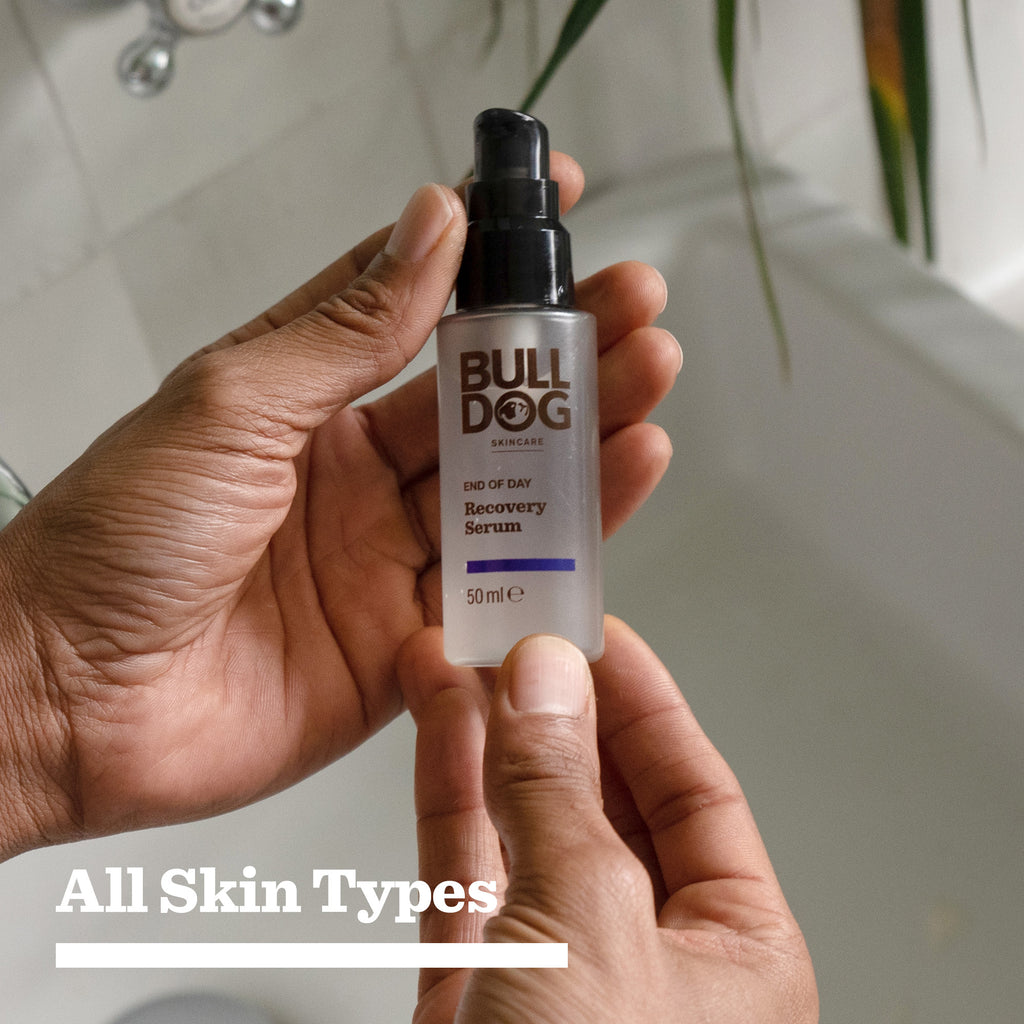 Someone holds a bottle from the End of Day Recovery Trio, showcasing its natural origin ingredients. A white bathtub is seen in the background. The bottom text states: All Skin Types.
